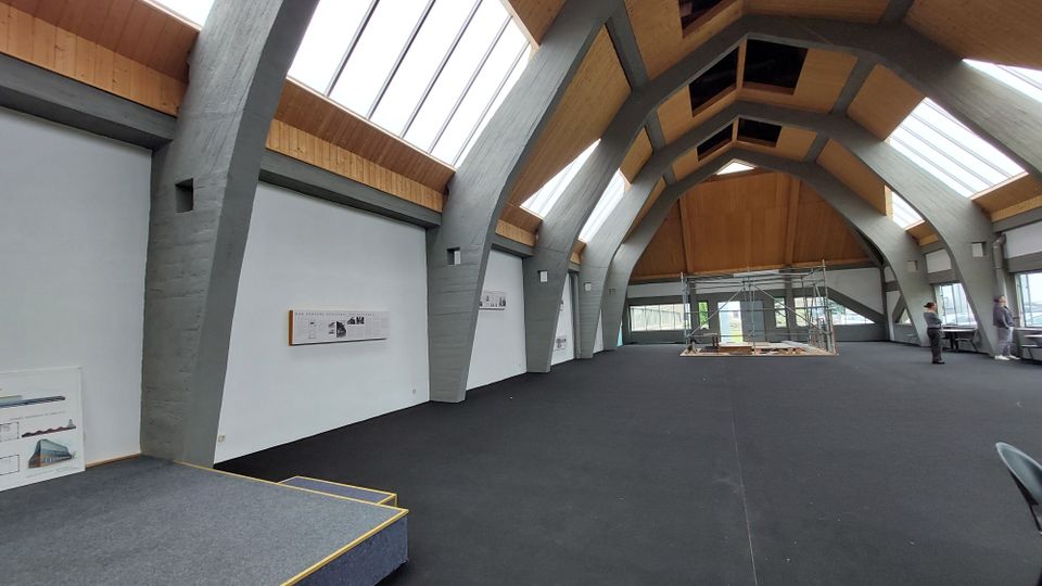 Innenraum eines modernen Gebäudes mit großen, bogenförmigen Betonstützen, einer Holzdecke und breiten Oberlichtern, die viel Tageslicht einlassen. Der Raum hat einen schwarzen Teppichboden, eine kleine Bühne links und eine Baustelle mit Gerüst in der Mitte.