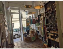 Kleiner Laden mit Holzfußboden und dekorativen Waren wie Schmuck, Kleidung und Karten. Im Vordergrund stehen Regale und ein Schaufenster mit Blick auf eine Straße und parkende Autos im Hintergrund.