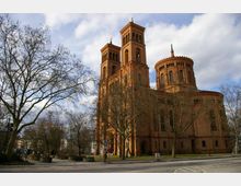 Historische Backsteinkirche mit zwei hohen Türmen und einer runden Apsis, umgeben von kahlen Bäumen. Die Kirche steht an einer Kreuzung, daneben befindet sich ein kleiner Platz mit Blumenbeeten.