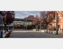 Bahnhofsvorplatz der S-Bahn-Station Wildau mit Kopfsteinpflaster, rotbraunen Bäumen und modernen Sitzbänken. Im Hintergrund sind das Glasdach des Bahnsteigs mit einer Bahnhofsuhr sowie ein Zug und ein rotes Backsteingebäude zu sehen.