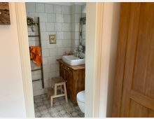 Badezimmer mit weißen Fliesen an den Wänden und gemusterten Fliesen auf dem Boden, ausgestattet mit einem Holzwaschtisch, einer weißen Aufsatzwaschschale, einem Spiegel mit verziertem Rahmen, einer Holzleiter als Handtuchhalter und einer kleinen Holztrittstufe.