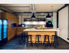 Moderne Kücheneinrichtung mit einer zentralen Kücheninsel aus hellem Holz und einer weißen Arbeitsplatte, umgeben von schwarzen Barhockern. Im Hintergrund befinden sich hölzerne Einbauschränke, ein Kühlschrank mit Glastüren, offene Regale mit Küchenutensilien, sowie eine Kaffeemaschine und eine Spüle; Metallrohre und Hängelampen betonen den industriellen Stil.