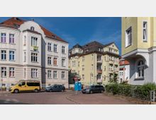 Straßenszene mit mehreren Altbau-Wohnhäusern, hell gestrichen in Beige- und Gelbtönen, mit roten Ziegeldächern und dekorativen Fassadenelementen. Im Vordergrund stehen parkende Autos, ein gelber Transporter, und ein Straßenschild an einer gepflasterten Kreuzung.