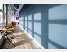 Langer, schmaler Flur mit Betonboden, einer Wand in Blau und einer Fensterfront, die Lichtmuster und Schatten auf die Wand wirft. Im Vordergrund steht ein kleiner Holztisch mit zwei Stühlen, im Hintergrund befinden sich bunte Sitzmöbel.