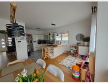 Offener Wohn- und Küchenbereich mit weißer Einbauküche und großer Kochinsel, darüber hängende Pendelleuchten und ein Fensterband an der Rückwand. Im Vordergrund steht ein Holzesstisch, rechts befindet sich eine Spielecke mit Teppich, niedrigem Regal und Spielzeug auf hellem Holzboden.