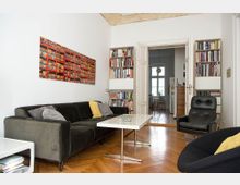 Wohnzimmer mit Parkettboden, schwarzem Sofa, gelben und grauen Kissen, modernem weißen Couchtisch und einem schwarzen Sessel. Im Hintergrund sind Bücherregale, eine geöffnete Doppeltür und eine Küche mit großen Fenstern sichtbar.