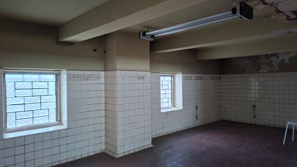 Ein leerer Raum mit weißen, abgenutzten Fliesenwänden bis zur halben Raumhöhe, zwei kleinen Fenstern mit Glasbausteinen und einer Decke mit sichtbaren Trägern und einer Leuchtstofflampe. Der Boden besteht aus dunklen, rötlichen Fliesen.