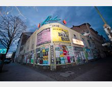 Eckgebäude eines Veranstaltungsortes mit dem Namen „Lido“, erkennbar durch eine leuchtende, blaue Neonaufschrift auf dem Dach. Die Fassade ist mit zahlreichen bunten Plakaten und Werbeanzeigen beklebt, während die Umgebung durch eine städtische Straßenecke mit kahlen Bäumen und Laternen bei Dämmerung geprägt ist.