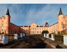 Innenhof eines schlossartigen, hellrosa Gebäudekomplexes mit zwei runden Ecktürmen und dunklen Spitzdächern sowie einer zentralen Fassade mit Zinnen. Ein breiter Weg führt durch den Hof, flankiert von niedrigen weißen Mauern mit Geländern, Sträuchern und kahler Vegetation unter blauem Himmel.