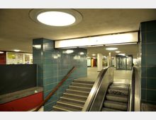 U-Bahn-Station mit einer Rolltreppe und Treppe, flankiert von Wänden mit blauen Fliesen. Die Beleuchtung besteht aus runden Deckenlampen, und im Hintergrund befinden sich verglaste Türen und Wände mit Werbeplakaten.