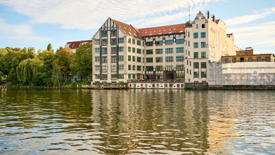 Großes mehrstöckiges Ufergebäude mit weißer Fassade, dunklen Fachwerk-Elementen und roten Ziegeldächern direkt am Fluss, mit vielen Fenstern und einem kleinen Anleger am Wasser. Links grenzt eine Baumreihe ans Ufer, rechts ist ein Gebäudeteil mit Baugerüst und Schutzplane zu sehen.