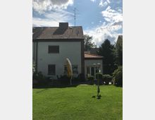 Zweistöckiges Einfamilienhaus mit heller Fassade und rotem Ziegeldach, im Vordergrund ein gepflegter Garten mit Rasenfläche, einem gelben Sonnenschirm und einer Laterne. Im Hintergrund sind Bäume und ein blauer Himmel mit Wolken zu sehen.
