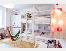 Helles Kinderzimmer mit einem Etagenbett aus Holz, einer Regenbogenhängematte und bunten Lichterketten an der Wand. Im Raum befinden sich außerdem ein weißer Schrank, Spielzeug und eine helle Holzböden.