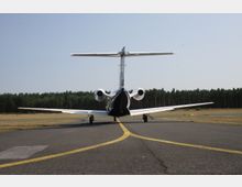 Ein kleines Passagierflugzeug steht auf einer asphaltierten Rollbahn, mit gelben Markierungslinien im Vordergrund. Im Hintergrund ist eine Waldlinie unter klarem Himmel zu sehen.
