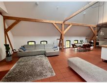 Ein offener Wohnraum in einem Dachgeschoss mit sichtbaren Holzbalken, weißen Wänden und Terrakottafliesen. Der Raum ist mit einer großen grauen Couch, einem Teppich, einem Esstisch mit Stühlen und dekorativen Pflanzen ausgestattet; mehrere kleine Fenster lassen Tageslicht herein.