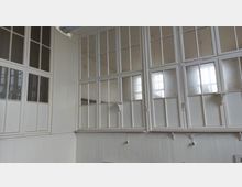 Innenraum mit weißen, großflächigen Fenstern im alten Holzrahmen-Stil, die den oberen Teil der Wand bedecken; darunter weiße Holzvertäfelungen und sichtbare Steckdosen.