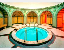 Runder Innenraum mit einem zentralen, türkisfarbenen Whirlpool, umgeben von in Mosaik gehaltenen Wänden und ornamentalen Bögen. Der Raum ist mit warmen Farben, dekorativen Details und einem Sitzbereich ausgestattet.