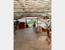 Ein Innenraum in einem wintergartenähnlichen Gebäude mit Wellstoffdach. Der Raum ist mit unterschiedlichen Möbeln eingerichtet, darunter Holztische, Stühle und eine Fass-Dekoration, mit Blick auf einen Garten durch große Glasfenster im Hintergrund.