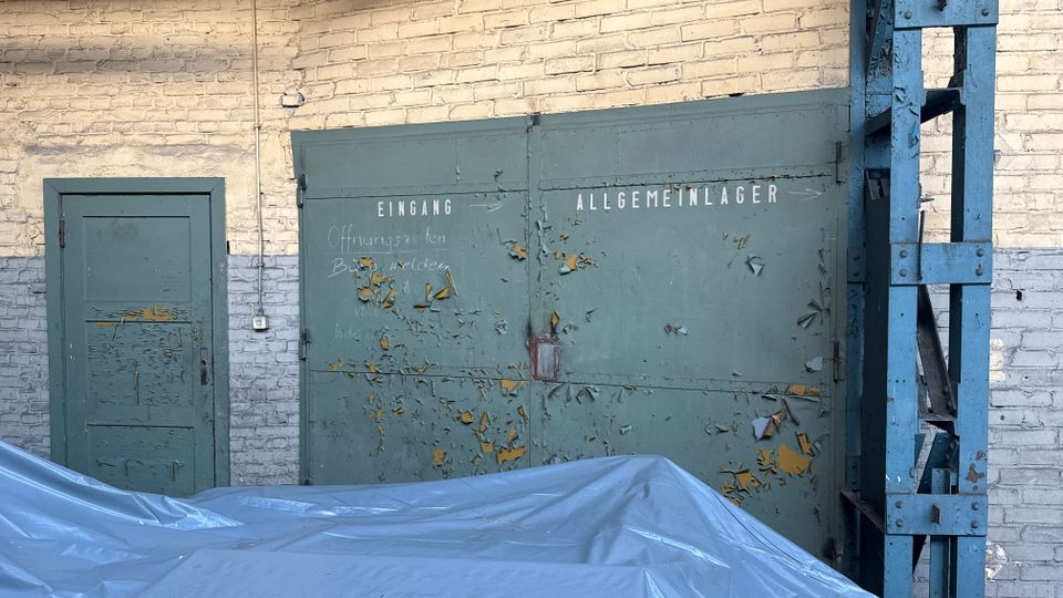 Außenwand eines Industriegebäudes aus weiß gestrichenem Ziegel mit einer kleinen Tür und einem großen, abblätternden grünen Doppeltor mit der Aufschrift „Eingang“ und „Allgemeinlager“. Rechts steht eine blaue Stahlkonstruktion, im Vordergrund liegt eine graue Abdeckplane.