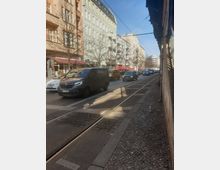 Straßenansicht in einer Stadt mit mehrstöckigen Wohnhäusern und Geschäften im Erdgeschoss. Im Vordergrund verlaufen Straßenbahnschienen neben einer Straße mit parkenden und fahrenden Autos; es ist eine ruhige urbane Umgebung bei Tageslicht.