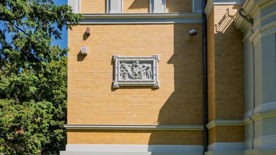 Seitenfassade eines gelblichen Ziegelgebäudes mit hellen Gesimsen und einer dekorativen Reliefplatte in weißem Rahmen. Links ragen hohe Bäume und Grün in das Bild, rechts sind Gebäudeecken und Fallrohre zu sehen.