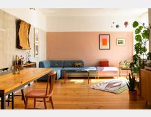 Ein Wohnzimmer mit Holzboden und modernem Interieur. Es umfasst eine blaue Ecksofa-Gruppe, einen Holztisch mit Stühlen, dekorative Pflanzen sowie bunte Wandbilder und Accessoires an einer rosa und naturfarben gestalteten Wand.