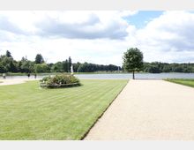 Weitläufiger Landschaftsgarten mit einem gepflegten Rasen, einem Blumenbeet mit einer Skulptur in der Mitte und einem einzelnen Baum rechts. Im Hintergrund ein großer See, umgeben von Bäumen, sowie ein Fußweg, der am Ufer entlangführt.