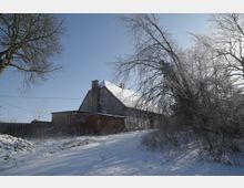 Winterliche ländliche Hofstelle mit einem großen, älteren Wohnhaus mit schneebedecktem Satteldach und einem niedrigen Backsteinanbau. Ein verschneiter Weg führt am Gebäude vorbei, umgeben von kahlen Bäumen und Sträuchern unter klarem Himmel.