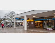 Außenansicht des U-Bahnhofs Britz-Süd in Berlin mit blauem Stationsschild. Links befinden sich ein Fahrradabstellbereich mit überdachten Stellplätzen, rechts ein Eingangsbereich mit Geschäften und Pflanzenverkauf.