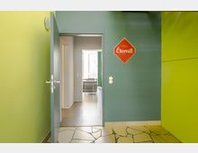 Ein Blick durch eine offene Tür in einem modernen Innenraum mit farbenfrohen Wänden in Grün- und Blautönen. Auf der blauen Wand hängt ein rotes Schild mit der Aufschrift "Eternit"; der Boden hat ein unregelmäßiges Steinmuster, und im hinteren Bereich ist ein Raum mit einem Sofa und großen Fenstern sichtbar.