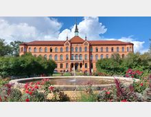 Historisches Backsteingebäude mit zahlreichen Rundbogenfenstern und einem zentralen Turm mit Uhr, umgeben von einem Garten mit Blumenbeeten, Büschen und einem Brunnen im Vordergrund. Der Himmel ist leicht bewölkt und blau.