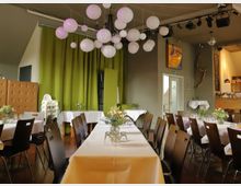 Ein gemütlicher Veranstaltungsraum mit gedeckten Tischen und Stühlen in Reihen angeordnet. Weiße Tischdecken, kleine Blumenarrangements als Dekoration, grüne Vorhänge im Hintergrund und weiße Papierlampions an der Decke bestimmen die Atmosphäre.