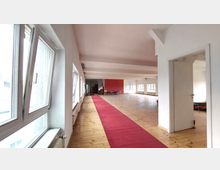 Geräumiger Innenraum mit Holzboden, weiß gestrichenen Wänden und mehreren großen Fenstern, die durch Tageslicht erhellt werden. In der Mitte des Raumes liegt ein roter Teppich, der zu einer roten Akzentwand im Hintergrund führt.