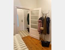 Ansicht eines Eingangsbereiches in einer Wohnung mit Holzdielenboden und einem anliegenden Flur mit gefliestem Boden im Schachbrettmuster. Im Vordergrund ist ein Kleiderständer mit Jacken und Taschen, links eine geöffnete Tür mit Sprossenglas, daneben ein Heizkörper.