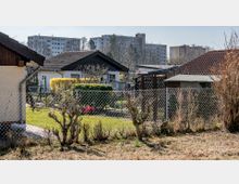 Kleingartensiedlung mit niedrigen Häusern, umgeben von Gärten und Maschendrahtzäunen; im Hintergrund sind mehrstöckige Wohnblöcke zu sehen. Die Szene zeigt eine Mischung aus urbanem und kleingärtnerischem Umfeld.