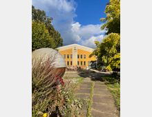 Außenansicht eines gelben Gebäudes mit rechteckigen Fenstern und einem Satteldach, umgeben von Bäumen und Pflanzen. Im Vordergrund führt ein mit Steinplatten befestigter Weg durch einen Garten, links ist eine runde Metallkonstruktion sichtbar.