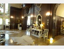 Eleganter Innenraum mit dunklen, holzvertäfelten Wänden, hohen Fenstern mit buntem Glas, heller Marmorboden mit Teppichen in Felloptik und einem dekorativen Kaminsims. Die Einrichtung besteht aus klassischen Möbeln, einem großen Wandspiegel und einem Hirschgeweih an der Wand.