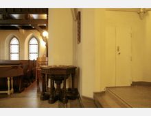 Innenraum einer Kirche mit Holzbänken, bunten Glasfenstern und einem kunstvoll geschnitzten, runden Taufbecken aus dunklem Holz. Im Hintergrund sind eine schlichte weiße Tür und ein beleuchteter Wandbereich zu sehen.