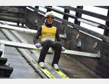 Ein Skispringer sitzt auf der Startrampe einer Sprungschanze, bereit zum Absprung, mit gelber Wettkampf-Weste und Helm. Die hölzerne Struktur der Rampe und Führungsschienen sind deutlich sichtbar.