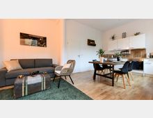 Wohnzimmer mit offener Küche: Ein graues Sofa und ein Korbsessel stehen auf einem grünen Teppich, davor eine alte Truhe als Couchtisch. Im Hintergrund eine kleine Küche mit weißen Hängeschränken, Holzarbeitsplatte und Esstisch aus Holz mit schwarzen Stühlen.