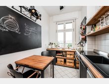 Kompakte, helle Wohnküche mit großem Fenster, Holzarbeitsflächen und einem kleinen Esstisch. Links hängt eine große Tafelwand mit Kreidezeichnungen, rechts sind offene Regale mit Gläsern, aufgehängten Töpfen sowie ein Herd und Spülbecken zu sehen.