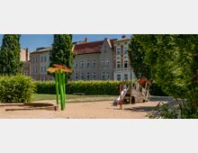 Städtischer Spielplatz mit Sandboden, einer Holzrutsche und bunten Spielgeräten in Form von Blumen, umgeben von Bäumen, Sträuchern und mehrstöckigen Wohnhäusern im Hintergrund.
