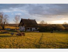 Fachwerkhaus mit Reetdach auf einer großen Wiese, umgeben von Bäumen und Buschwerk im ländlichen Gebiet, unter einem teils bewölkten Himmel bei Sonnenuntergang. Im Vordergrund befinden sich eine Holzkarre und gestapeltes Brennholz.
