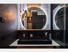 Modernes Badezimmer mit dunklen Fliesen, schwarzem Waschbecken und hölzernem Unterschrank. Ein runder, hinterleuchteter Spiegel dominiert die Szene, ergänzt durch hängende Glühbirnen und minimalistische Dekorationen.