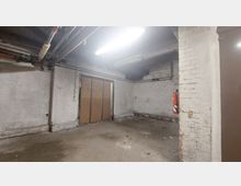 Leerer Kellerraum mit unverputzten, weißen Ziegelwänden, einem Boden aus Beton und sichtbaren Rohren an der Decke. Im Raum befinden sich doppelte Holztüren und ein an der Wand montierter Feuerlöscher.