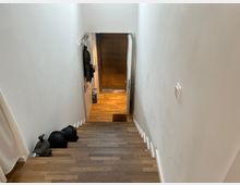 Holztreppe in einem Innenraum, die zu einem Flur mit Parkettboden führt. An der rechten Wand befindet sich ein Lichtschalter, und links an der Wand hängt ein Kleidungsstück neben einer angelehnten Tür.