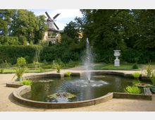 Ein gepflegter Garten mit einem runden Wasserbecken, in dem ein Springbrunnen sprudelt und Seerosen schwimmen. Im Hintergrund sind dichte Bäume, eine historische Windmühle und ein dekorativer Steinpflanztrog sichtbar.