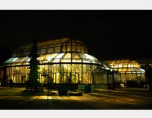 Ein großes, hell erleuchtetes Gewächshaus mit gewölbtem Glasdach und metallischen Strukturen, umgeben von einem dunklen Nachthimmel. Im Vordergrund befinden sich Bänke, gepflegte Grünflächen und ein einzelner Baum.
