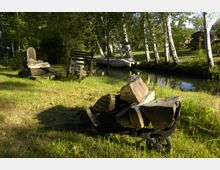 Ein ländlicher Garten mit einem kleinen Kanal im Hintergrund, flankiert von Birkenbäumen. Im Vordergrund liegen Holzscheite auf einer einfachen Schubkarre, daneben stehen eine alte Holzbank und ein kleiner handgezogener Wagen.