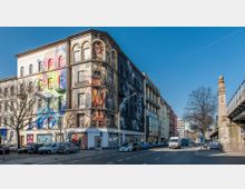 Mehrstöckiges Gebäude mit auffälligen Wandgraffitis in einer städtischen Straße; links Bäume ohne Laub und geparkte Autos, rechts ein erhöht verlaufendes Eisenbahnviadukt aus Stein. Moderner Stadtteil mit Mischarchitektur bei klarem Himmel.